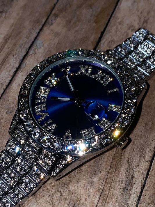 Silver-tone women's watch with a diamond-studded bezel and Roman numeral markers on a blue dial. The watch features a date display at the 3 o'clock position and a sparkling, crystal-encrusted band for a sophisticated, luxurious look.