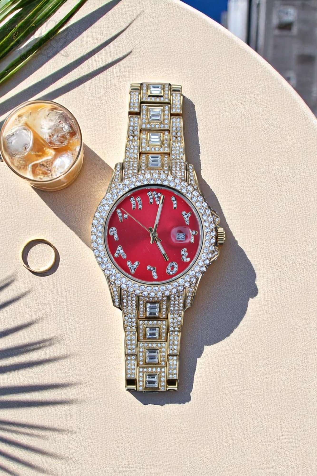Gold-tone unisex watch with a red dial, crystal-encrusted bezel, and rectangular stones on the band. The watch features sparkling Roman numeral markers and a date display, offering a luxurious and eye-catching design.